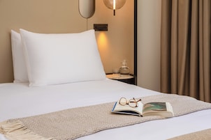 A neatly made bed with white linens, a pair of glasses, and an open book on a beige textured throw.