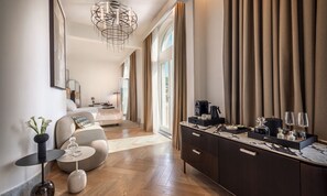 Ein modernes Hotelzimmer mit Bett, einer Sitzecke mit Sofa und Sesseln, einem Esstisch mit Kaffeemaschine und einem großen Fenster mit Vorhängen.