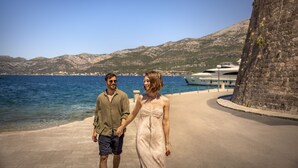 Um casal caminhando em um calçadão à beira-mar, com vista desobstruída para o mar e montanhas ao fundo.