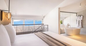 Une chambre moderne avec un grand lit, une vue sur la mer et une salle de bain séparée dotée d’une baignoire sur pieds.