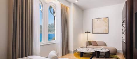 Habitación de hotel con cama, sofá, una mesita y vistas al mar a través de amplias ventanas.
