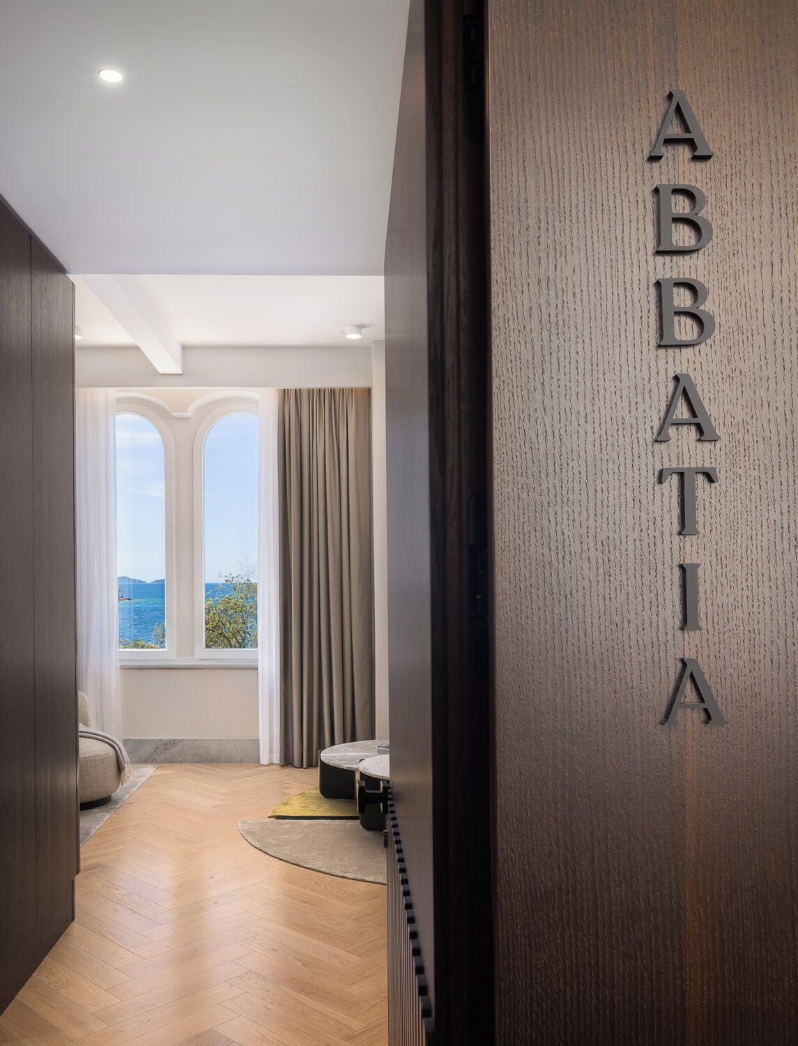 A hallway with a wooden floor, a view of the sea through a window, and a wall with the letters 'ABBATIA' on it.