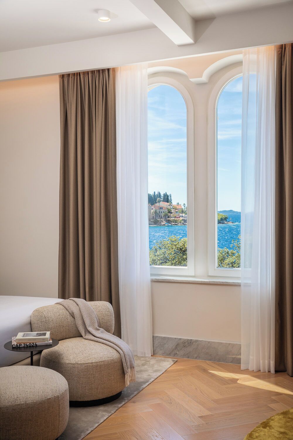 Un dormitorio con cama, dos otomanas, una mesita con un libro y vistas al mar a través de ventanas arqueadas.