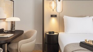 Habitación de hotel con cama, escritorio con lámpara, silla, mesita de noche redonda con lámpara y un tablero de ajedrez.