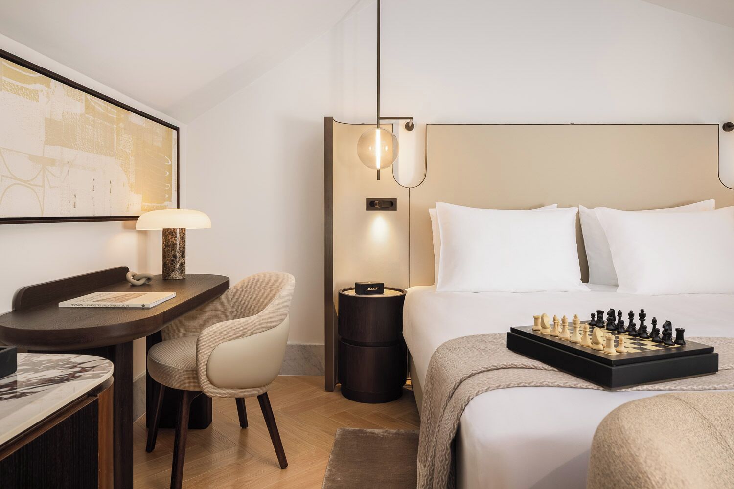 Habitación de hotel con cama, escritorio con lámpara, silla, mesita de noche y un tablero de ajedrez.