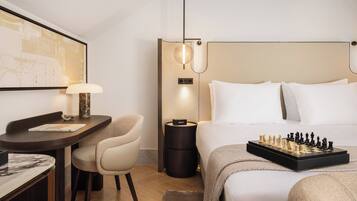 Habitación de hotel con cama, escritorio con lámpara, silla, mesita de noche y un tablero de ajedrez.
