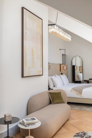 Ein modernes Hotelzimmer mit Bett, einer Sitzecke, einem kleinen Tisch mit einem Buch, einem gerahmten Kunstwerk und einer Kristallleuchter.
