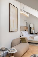 A modern hotel room with a bed, a sofa, a small table with a book, a framed artwork, and a chandelier.