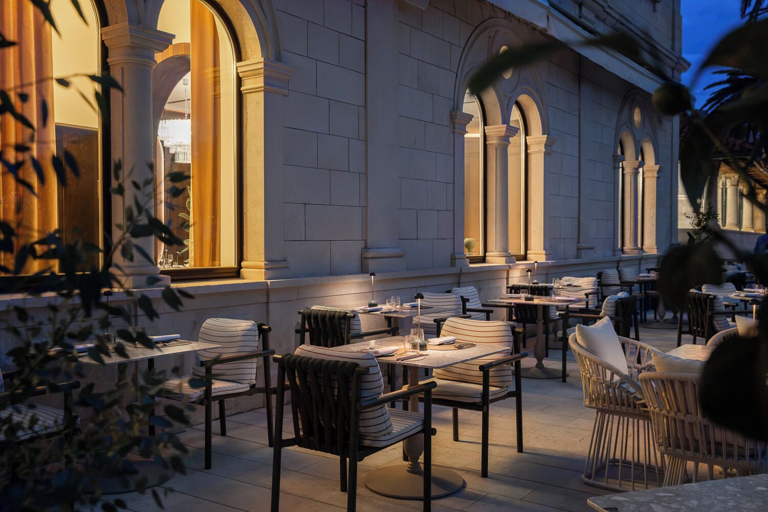 An outdoor dining area with tables and chairs, surrounded by a building with arched windows and columns.
