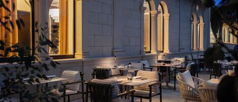 Un área de comedor al aire libre con mesas y sillas, rodeada por un edificio con ventanas arqueadas y columnas.