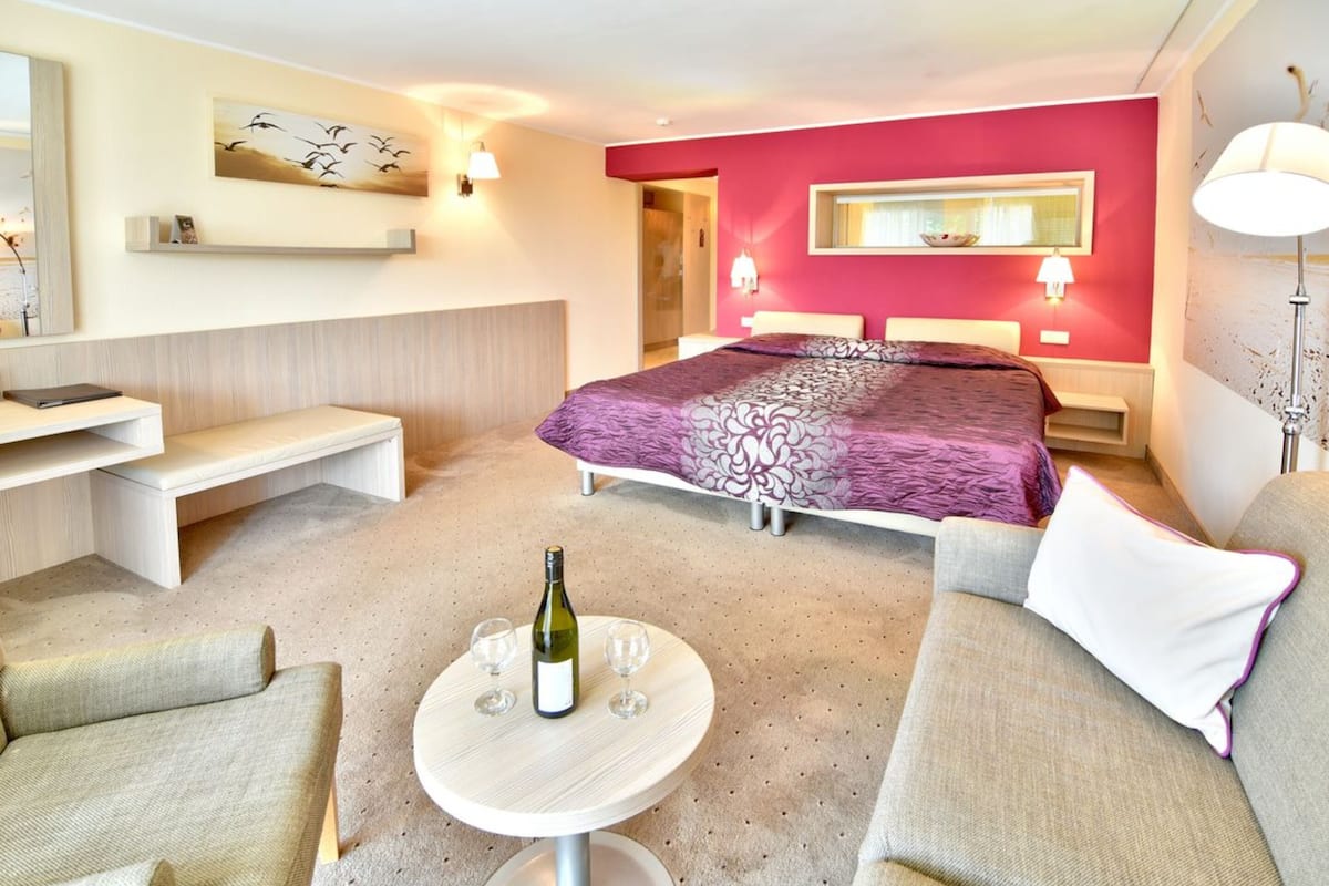 Una habitación de hotel moderna con una cama grande, una mesa de centro redonda, una botella y vasos, un sofá y un banco.