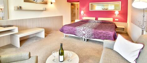 A modern hotel room with a large bed, a round coffee table with a bottle and glasses, a sofa, and a bench.