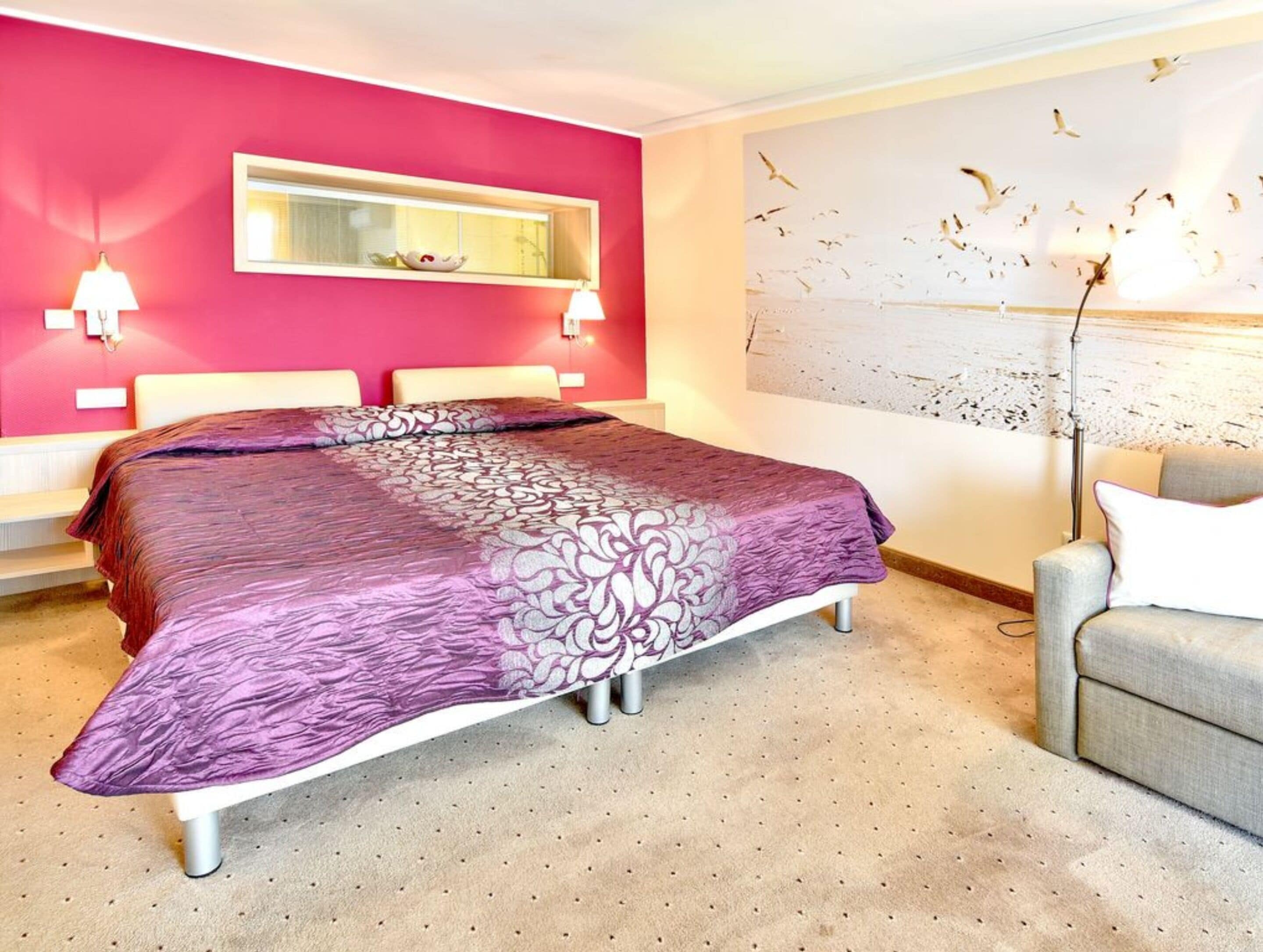 A bedroom with a bed, a window, a wall-mounted light, a decorative wall piece, and a chair.