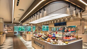 A modern hotel lobby with a central buffet area, a reception desk, and a seating area with a view of the outdoors.