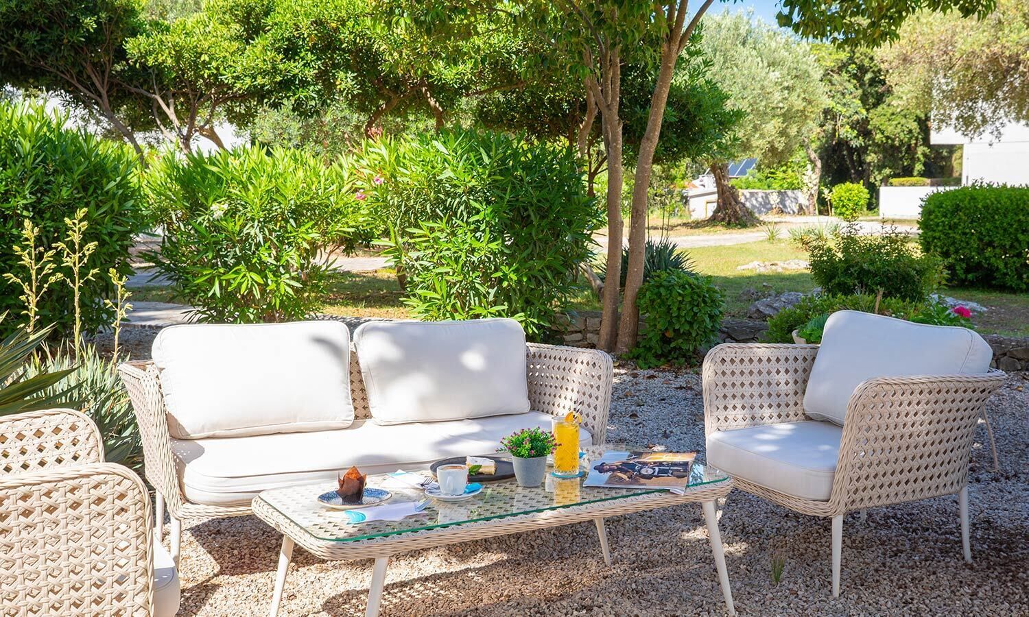 Un espace extérieur aménagé avec des meubles en osier et une table en verre, entouré de verdure et d’un mur de pierre.