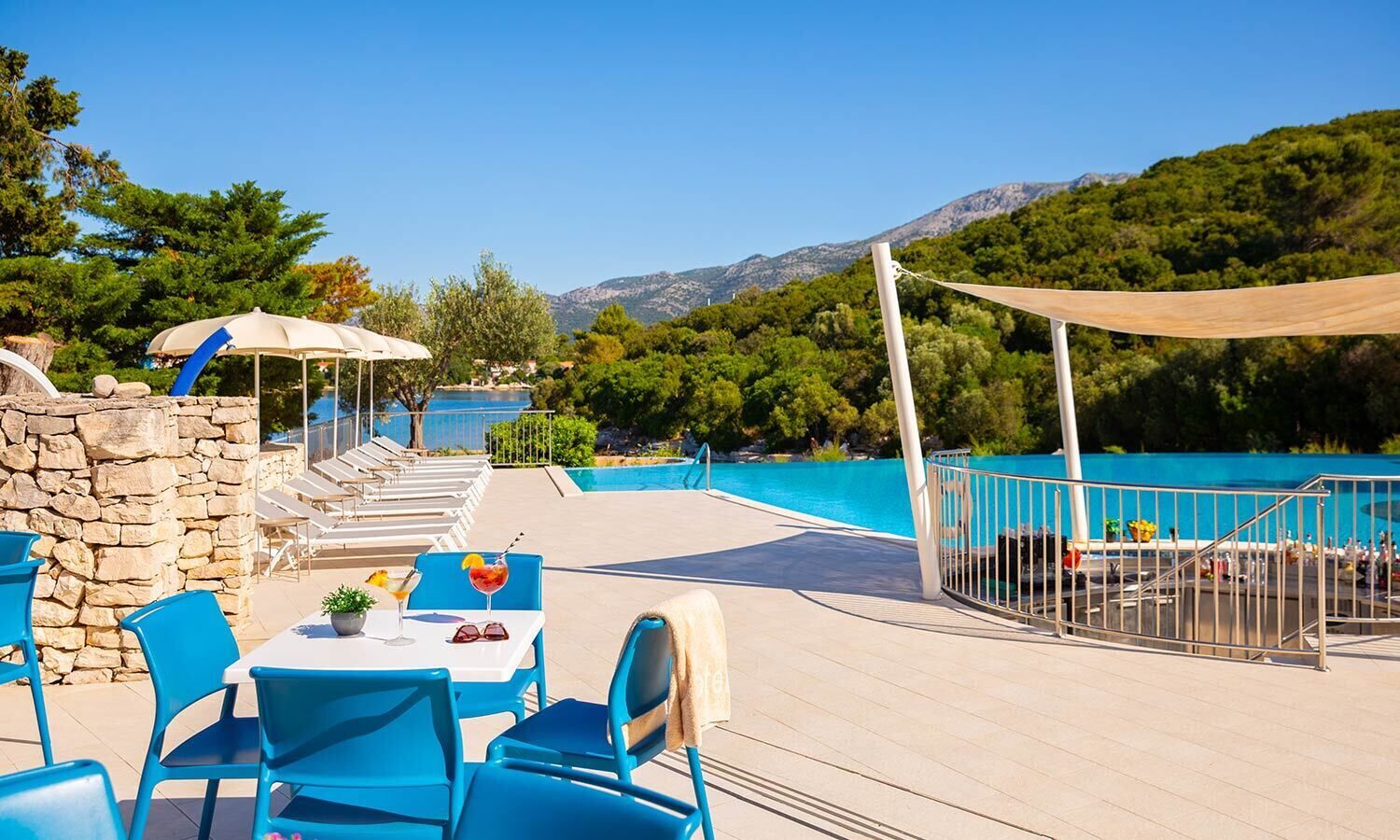 Un espace aménagé au bord de la piscine, avec des chaises bleues et une table, un mur en pierre, et une vue sur une montagne et l’eau.