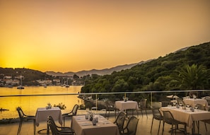 An outdoor dining area with tables and chairs, overlooking a body of water and a hilly landscape during sunset.
