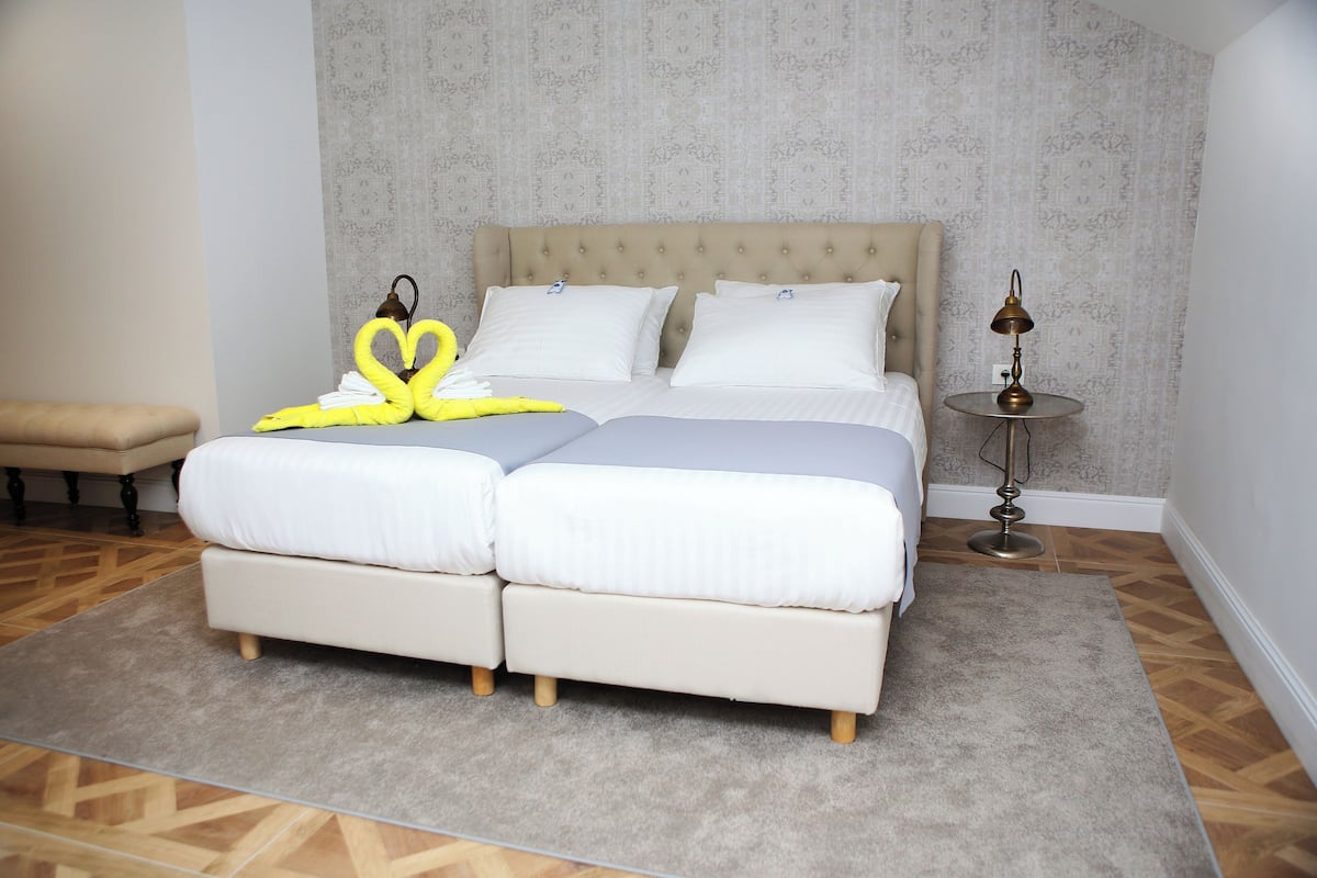 Uma cama de casal com cabeceira bege, uma mesa de cabeceira com abajur e um banco em um quarto com papel de parede estampado e piso de madeira.