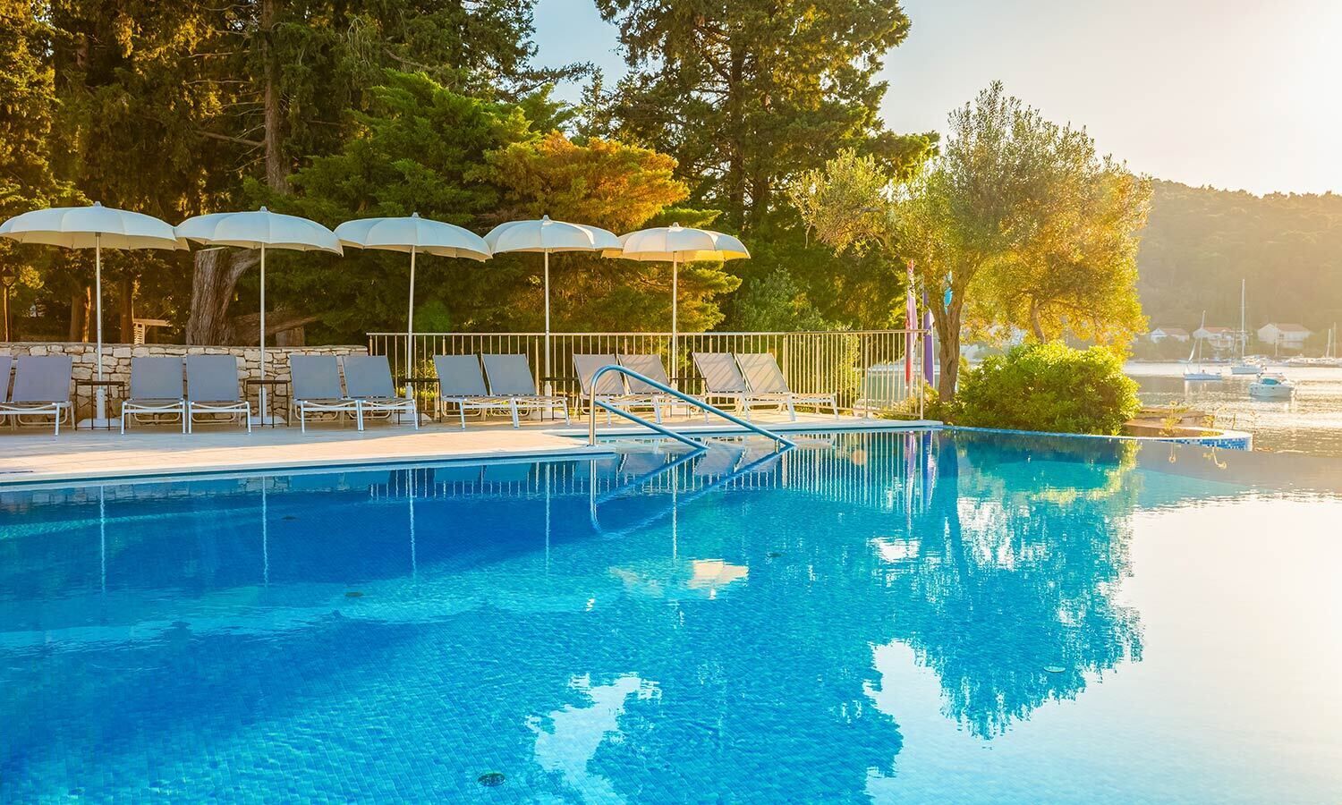 A swimming pool with lounge chairs and umbrellas, surrounded by trees and a view of boats.