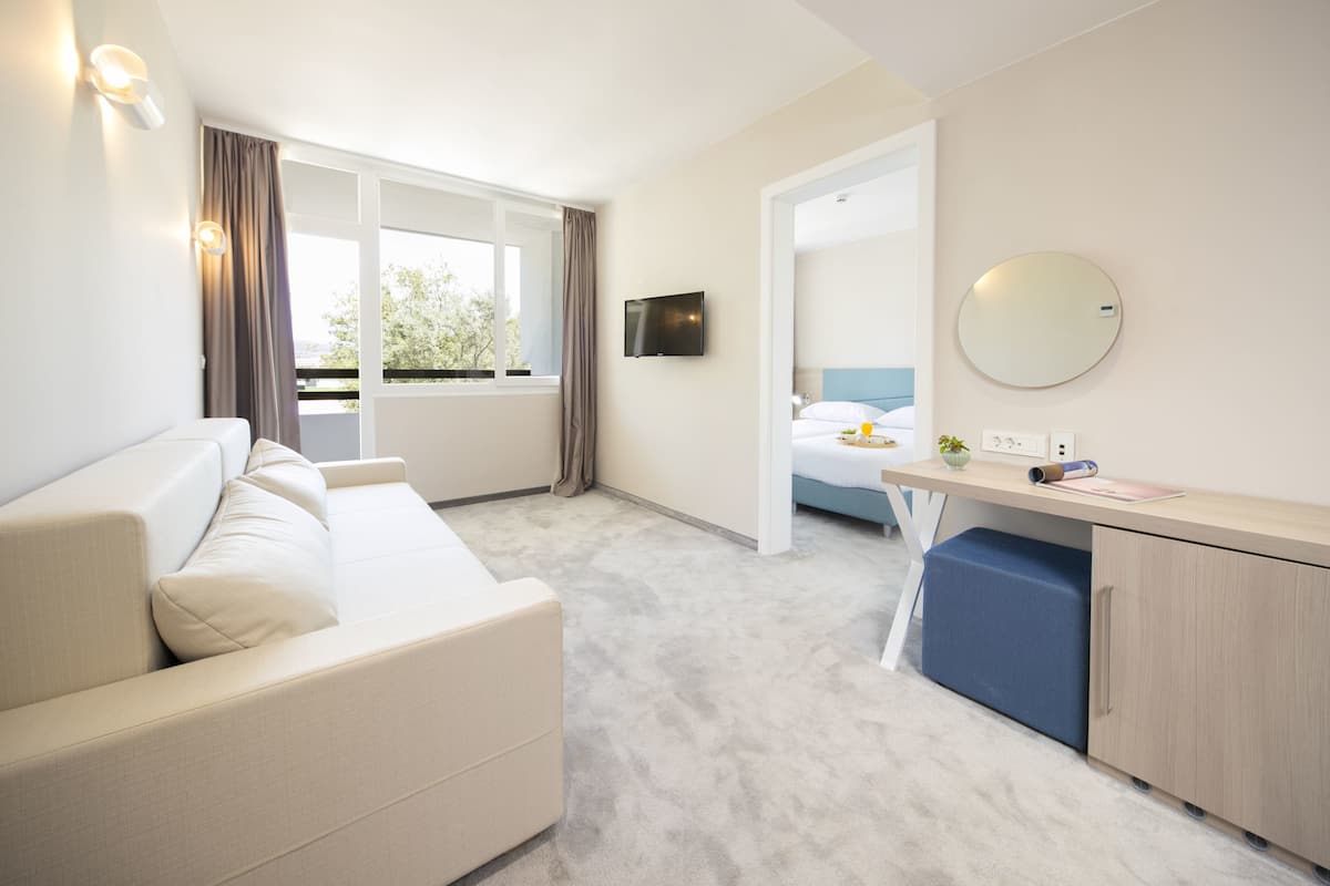 Quarto de hotel moderno com sofá, escrivaninha, cama e uma janela grande com cortinas.