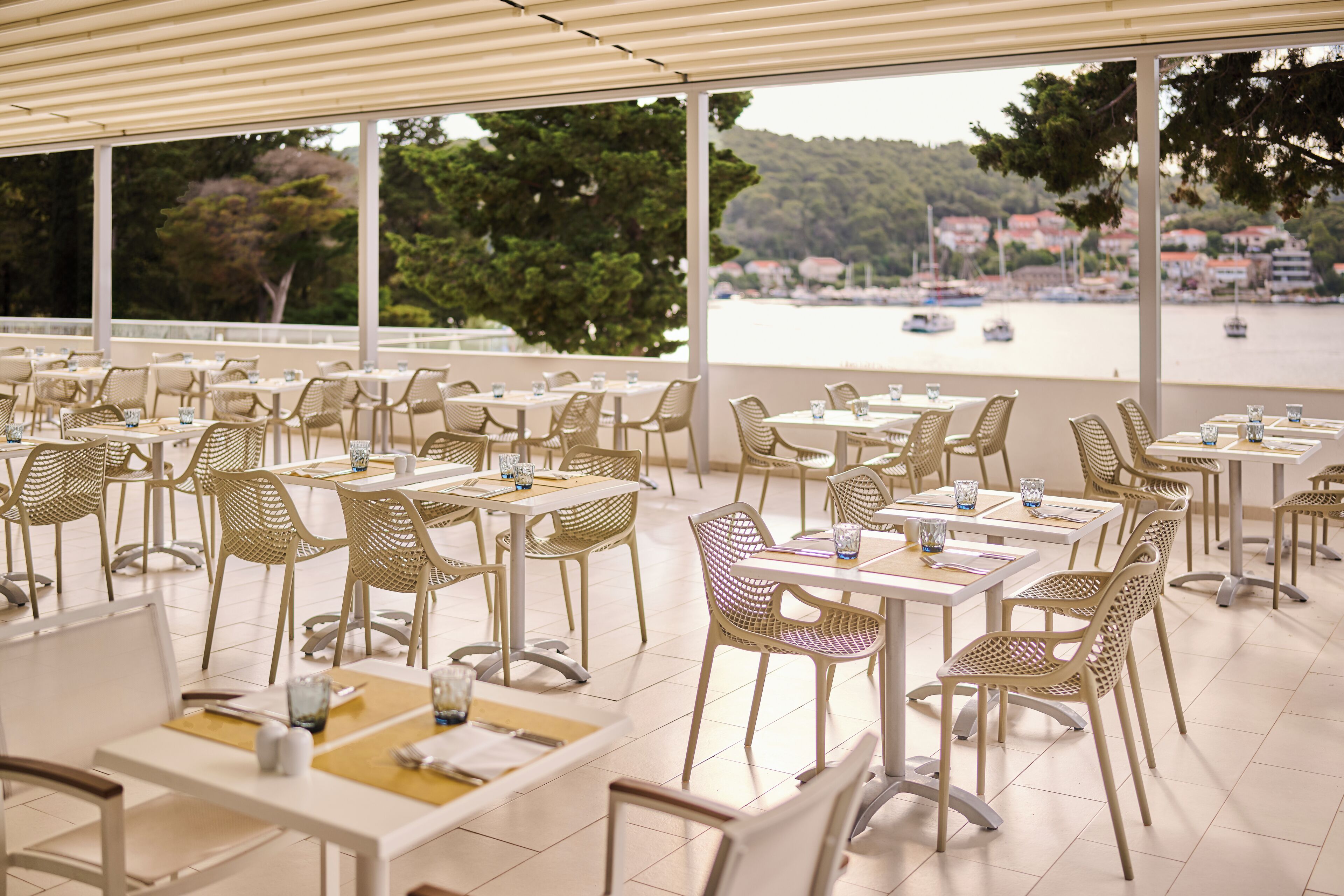A restaurant with outdoor seating overlooking a body of water and a town. Tables are arranged with wicker chairs and white tablecloths.
