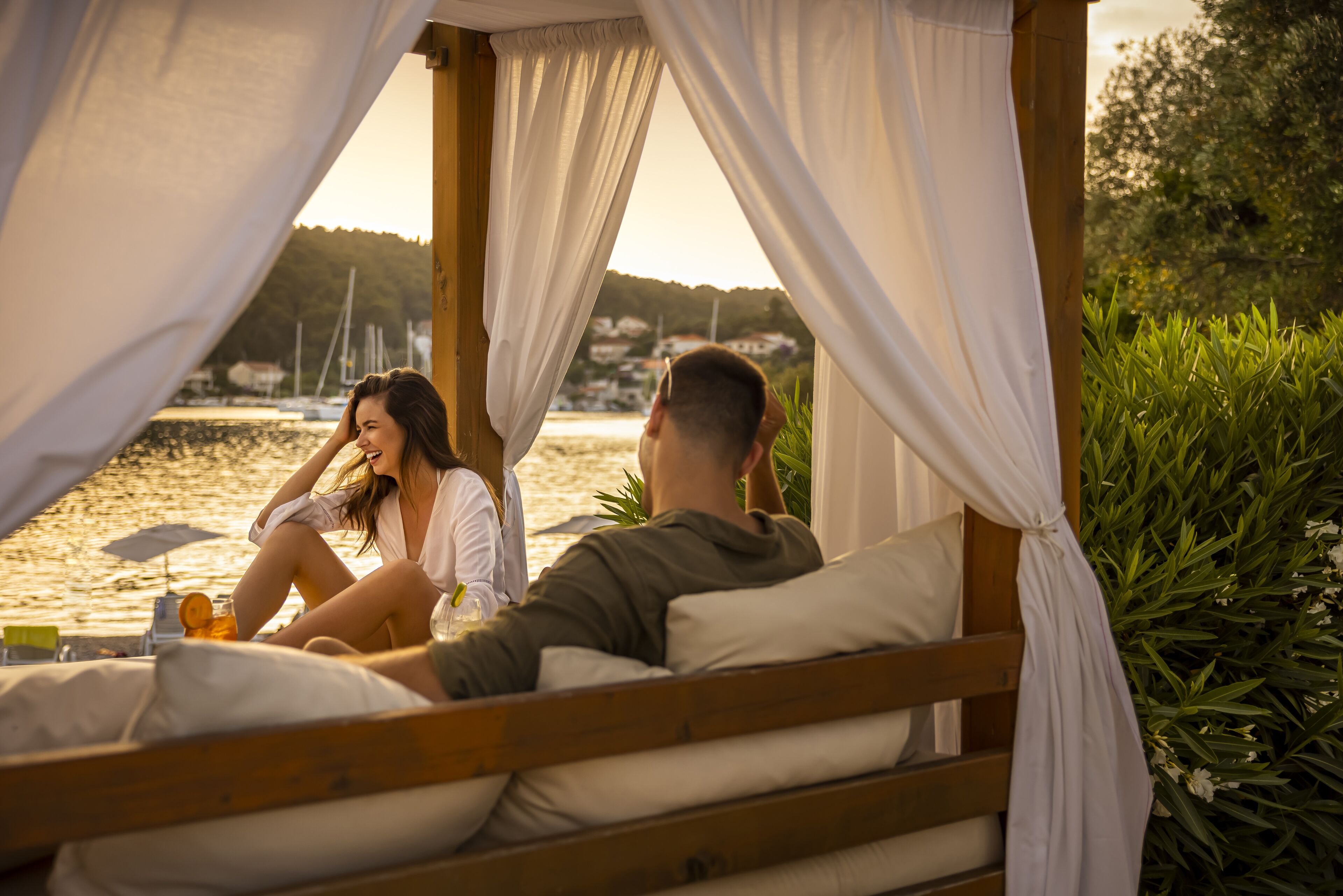 A couple relaxing on a wooden bed with white curtains, overlooking a body of water and greenery.