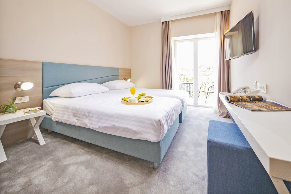 Quarto de hotel com cama, mesa de cabeceira, luminária e escrivaninha com cadeira.