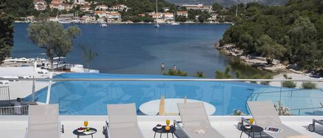Zona de piscina con sillones reclinables y una mesa con fruta y una bebida, con vistas a un lago con barcos y casas al fondo.
