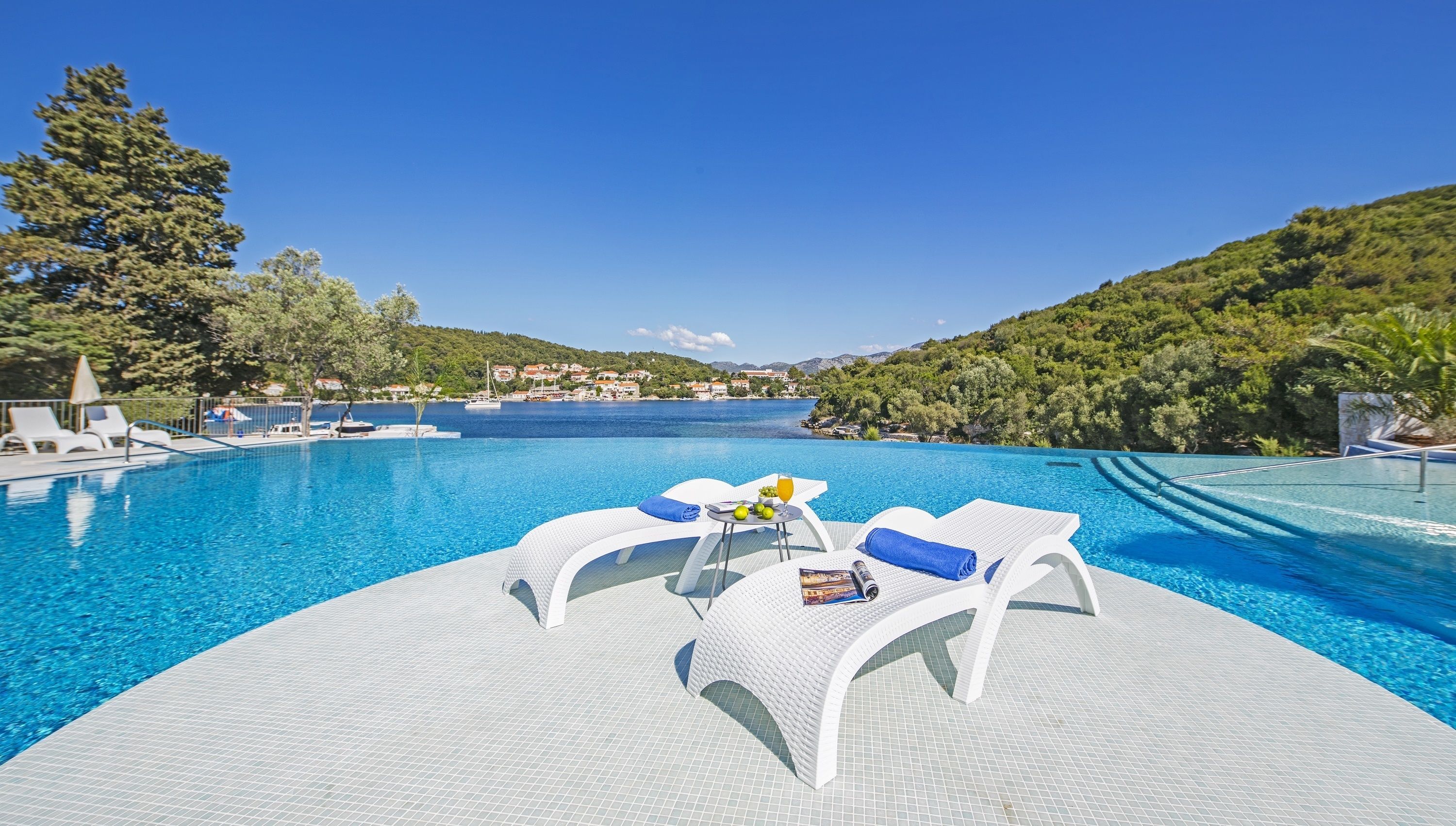 A poolside area with lounge chairs, a table with fruits and a drink, and a view of the sea and hills.