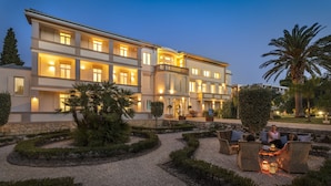 A multi-story hotel with a well-lit exterior, a landscaped garden with hedges and trees, and outdoor seating areas.