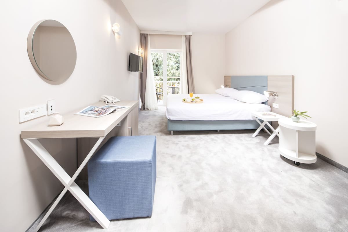 Quarto de hotel moderno com cama, escrivaninha, cadeira, mesa de cabeceira, espelho e uma pequena planta em vaso.