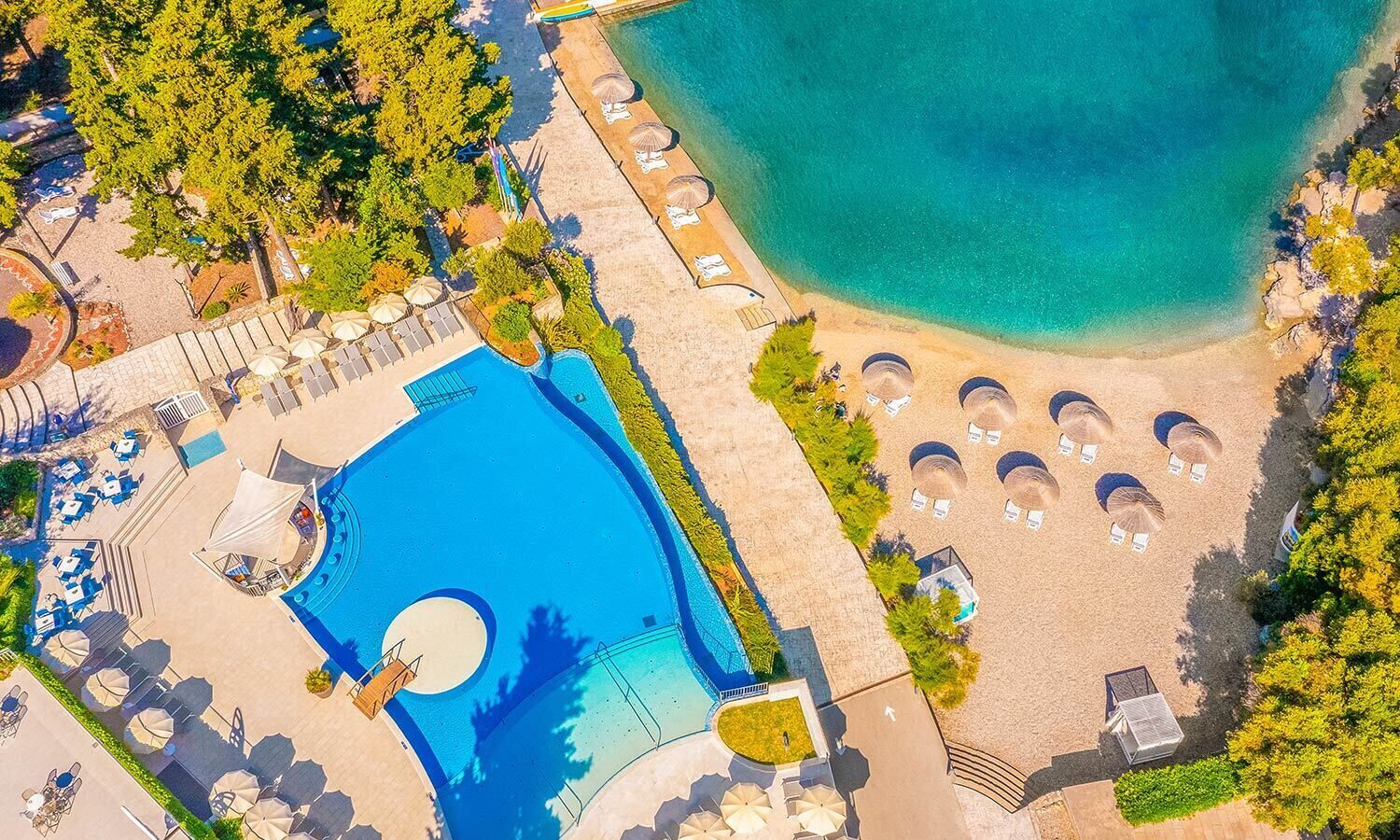 An aerial view of a resort with a large swimming pool, sun loungers, and a beach area.