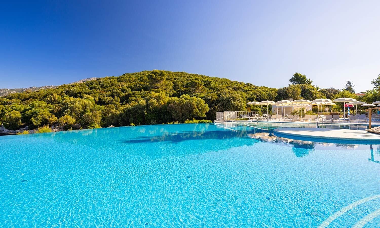 A clear blue pool with lounge chairs and umbrellas, surrounded by lush greenery and a mountain backdrop.