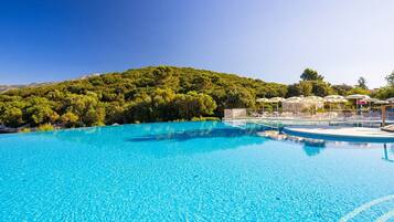A clear blue pool with lounge chairs and umbrellas, surrounded by lush greenery and a mountain backdrop.