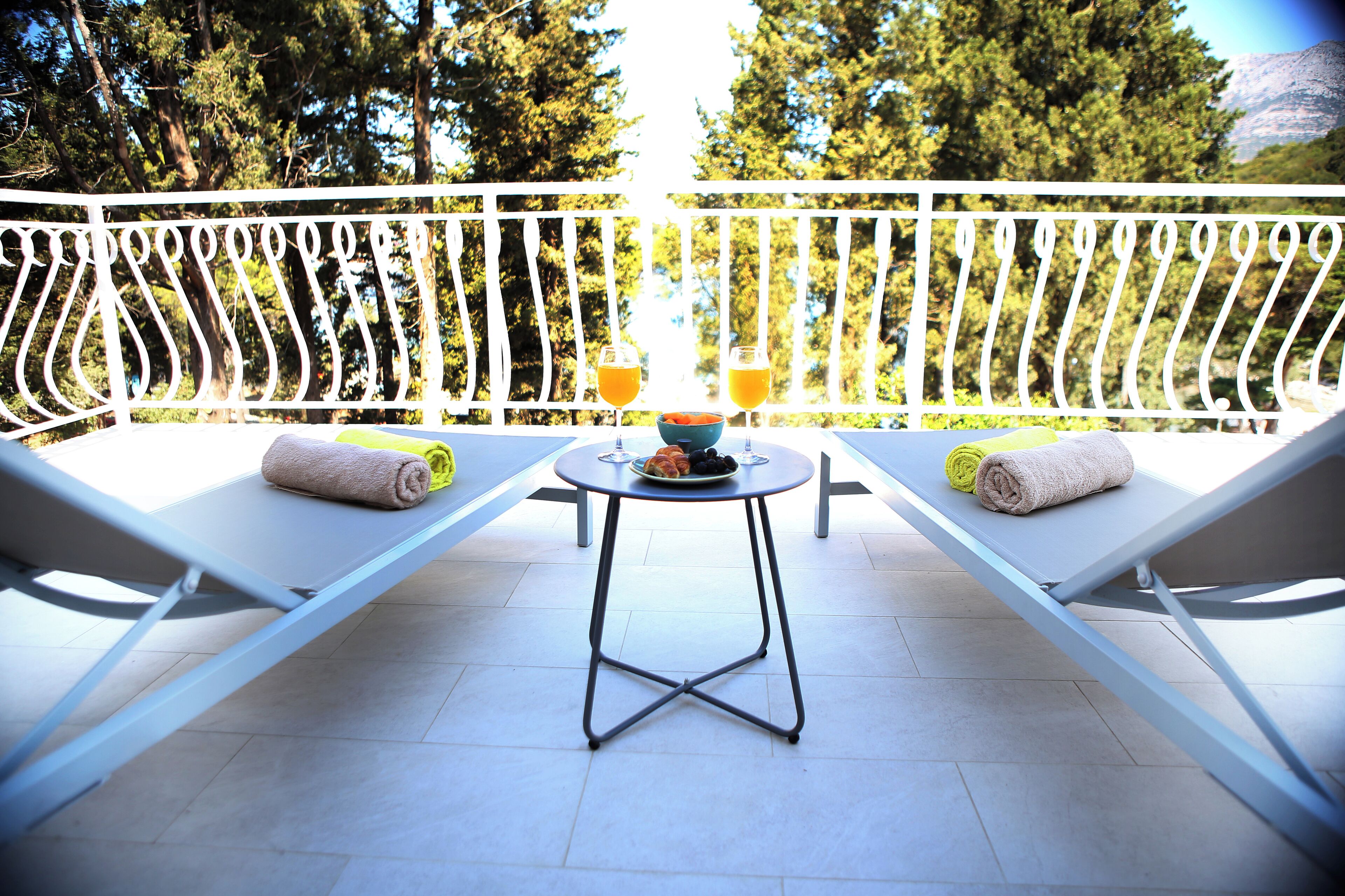 Un balcon avec deux fauteuils, une petite table sur laquelle se trouve un saladier rempli de fruits et deux verres de jus d’orange, ainsi qu’une balustrade blanche.