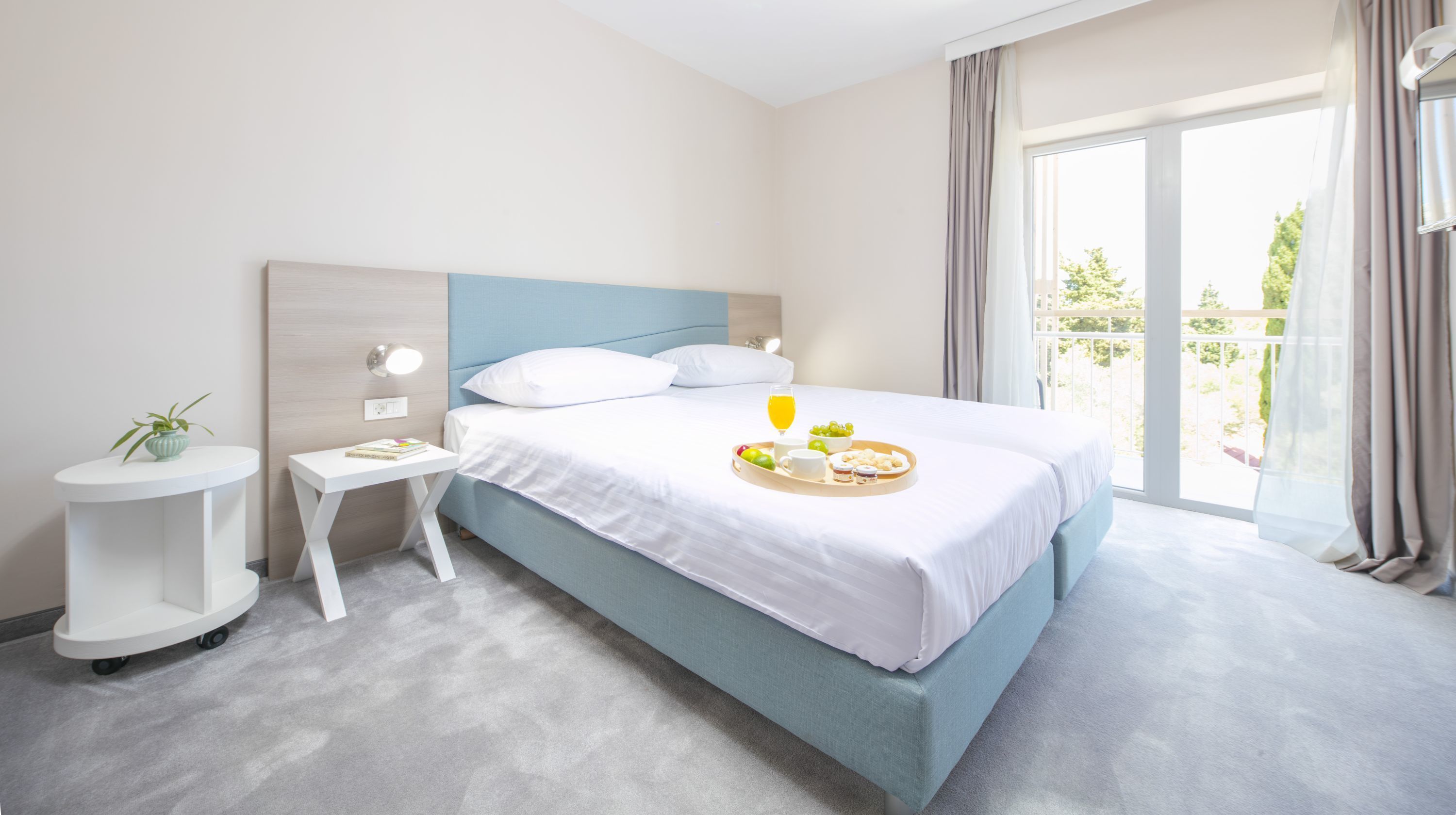 A hotel room with a bed, a bedside table, a small table with a plant, and a window with curtains.