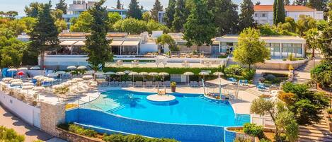 Ein großer Pool im Freien, umgeben von Liegestühlen und Sonnenschirmen, mit Blick auf Wohnhäuser und Bäume im Hintergrund.