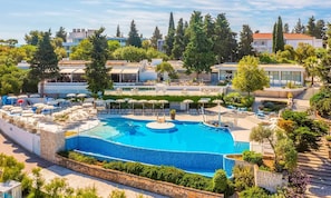 Ein großer Pool im Freien, umgeben von Liegestühlen und Sonnenschirmen, mit Blick auf Wohnhäuser und Bäume im Hintergrund.