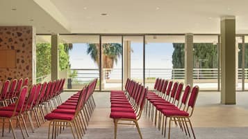 A conference room with rows of red chairs and a large glass window offering a view of palm trees and a balcony.