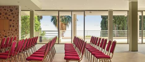 A conference room with rows of red chairs and a large glass window offering a view of palm trees and a balcony.