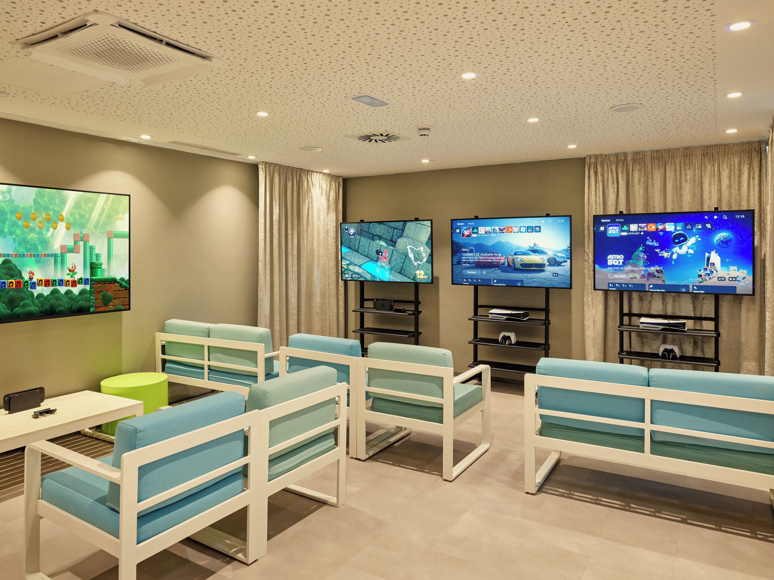 A modern hotel room with a gaming area featuring two large flat-screen TVs, comfortable seating, and a small table with a phone and remote control.