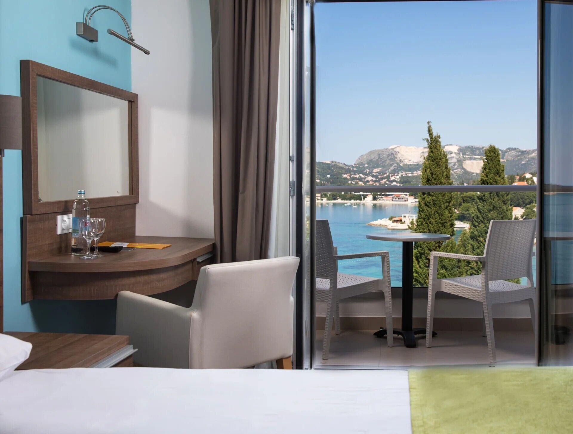 A hotel room with a bed, a desk with a mirror, a chair, a balcony with a table and chairs, and a view of the sea and mountains.