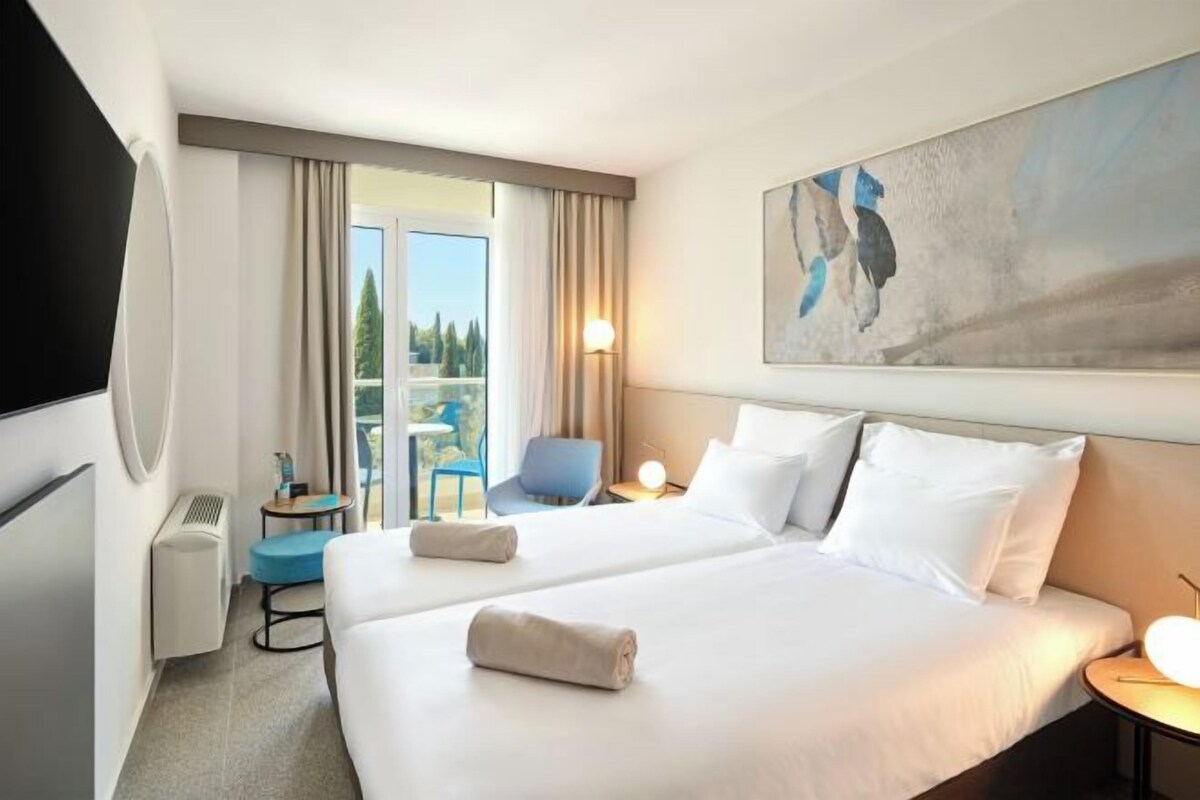 A hotel room with two beds, a chair, a desk, and a balcony with curtains.