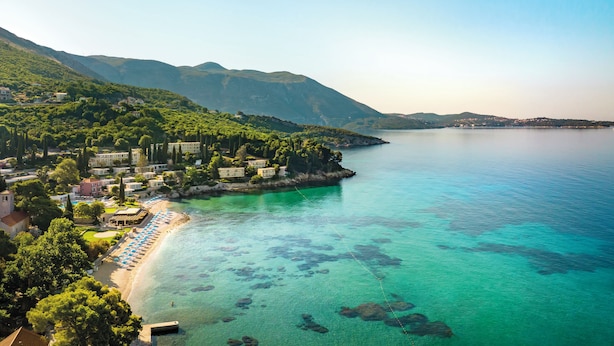 Un resort costero con playa, aguas turquesas cristalinas y montañas al fondo.
