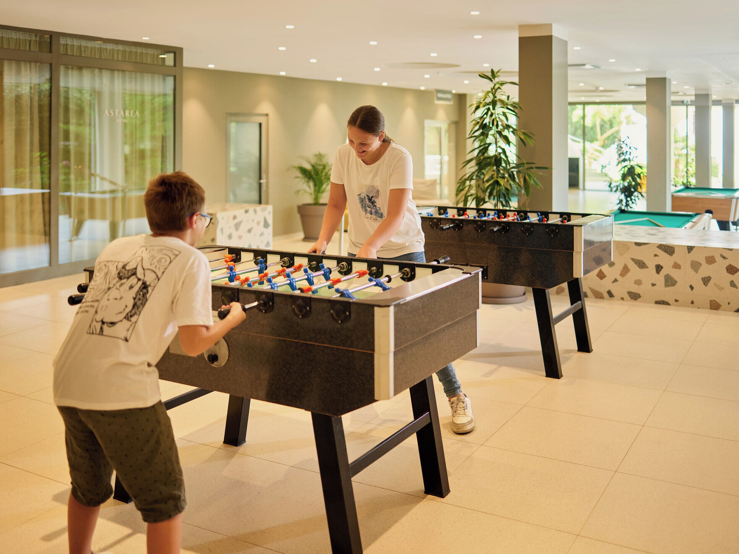 A modern indoor setting with two foosball tables, a pool table, and a potted plant.