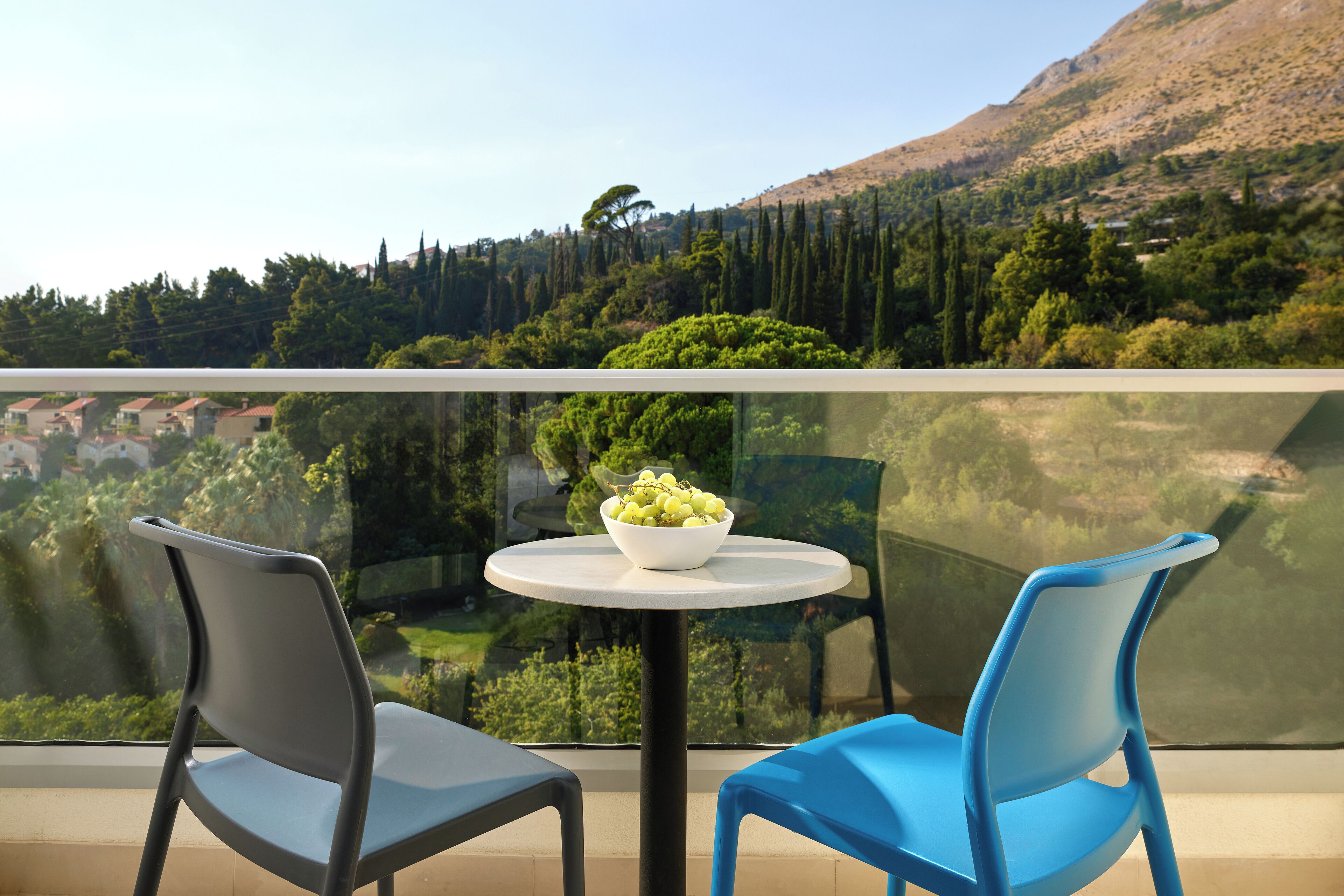 Ein Balkon mit zwei Stühlen und einem Tisch, einer Obstschale und einem schönen Ausblick auf eine Hügelslandschaft mit Bäumen und Gebäuden.