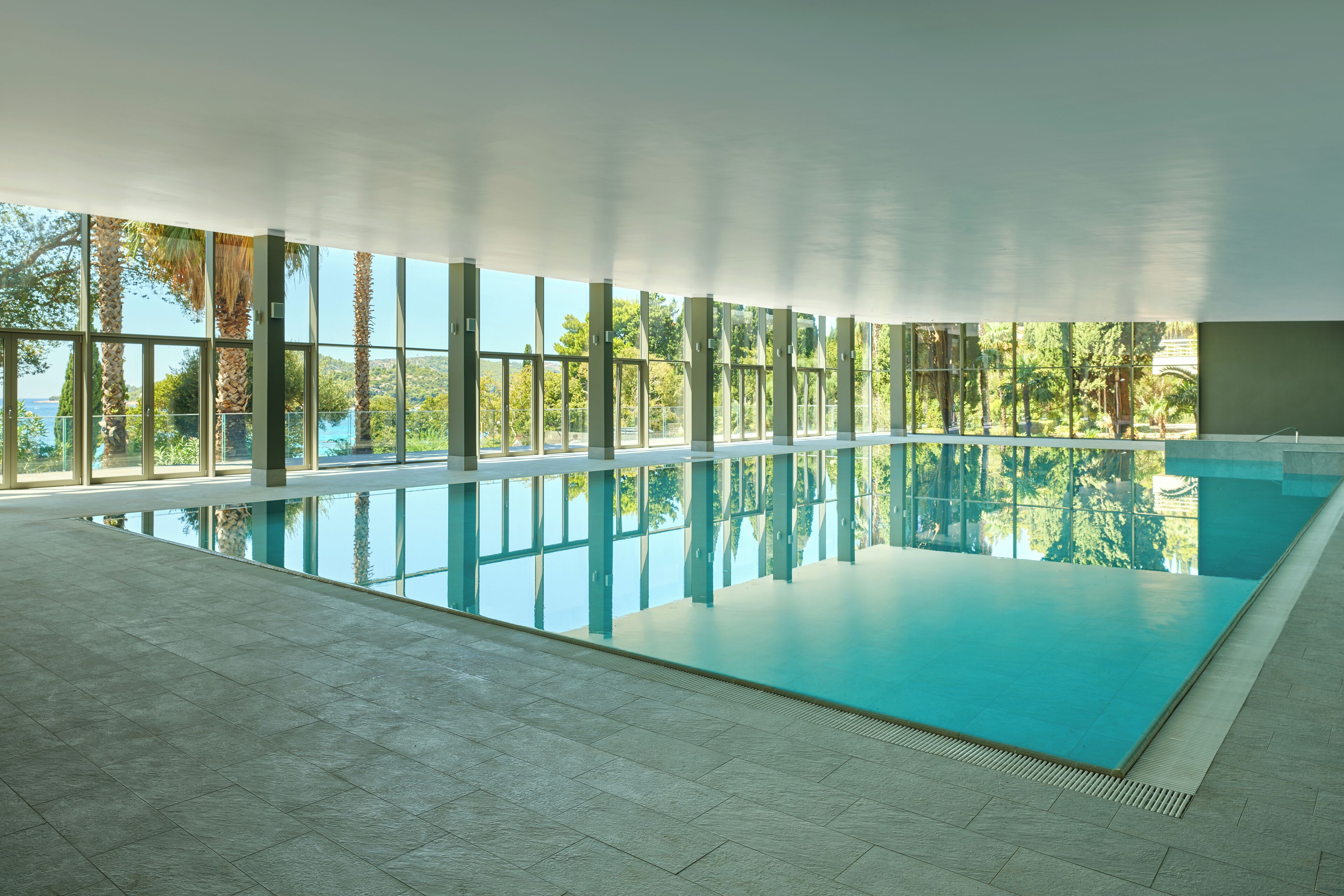 A large indoor swimming pool with clear glass walls, surrounded by a tiled floor, and a view of palm trees and greenery outside.