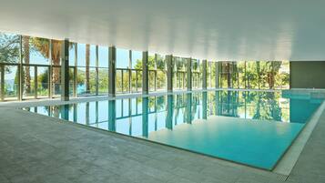 A large indoor swimming pool with clear glass walls, surrounded by a tiled floor, and a view of palm trees and greenery outside.