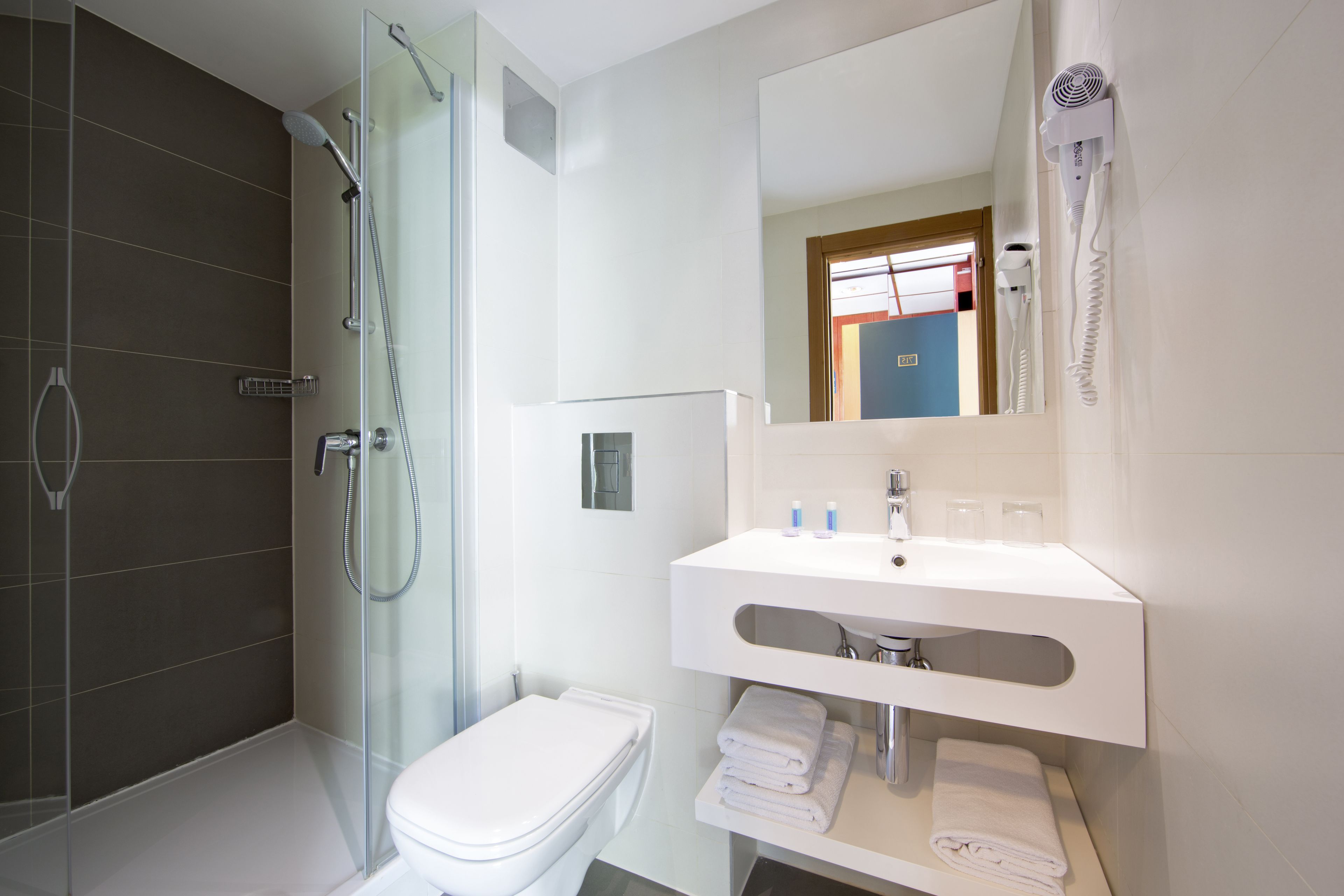 A modern bathroom with a glass-enclosed shower, a white toilet, a white sink with a mirror above it, and a hairdryer mounted on the wall.