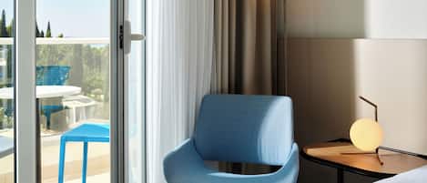 Quarto de hotel com uma grande porta de correr de vidro que dá para uma área externa com uma cadeira azul e uma mesa. Há uma cama com roupa de cama branca e uma mesa de cabeceira com um abajur.
